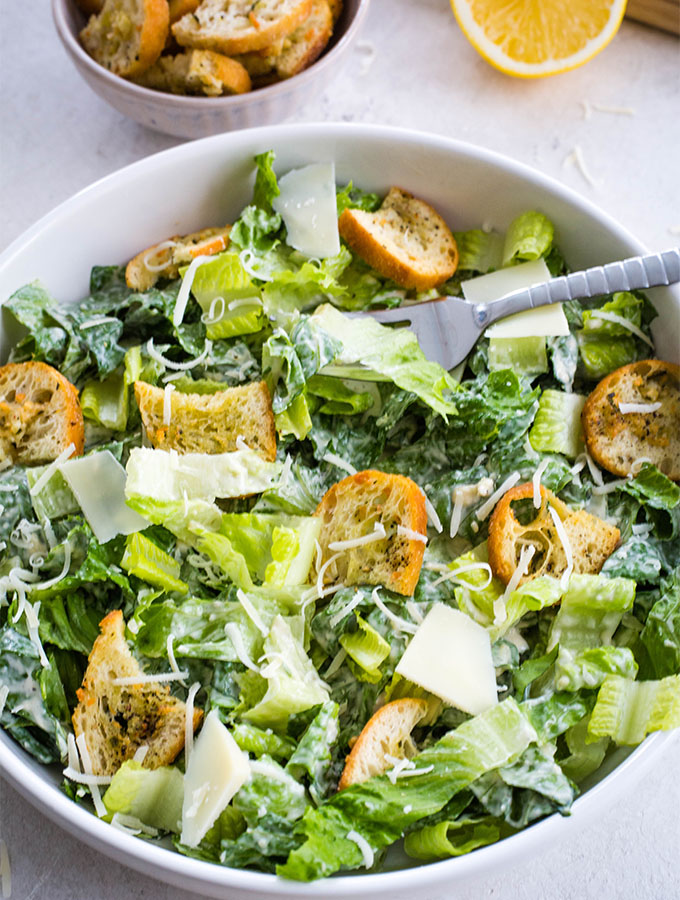 Caesar salad is plated in a white bowl with a fork and topped with shredded parmesan cheese.