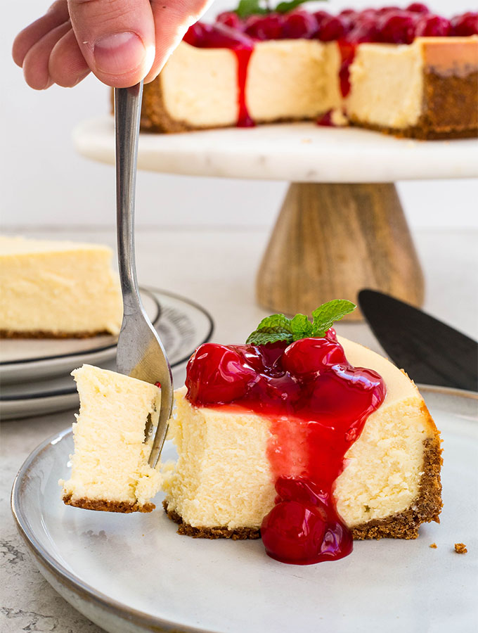 A fork is pulling a bite away from the slice of cheesecake to show fluffy texture.