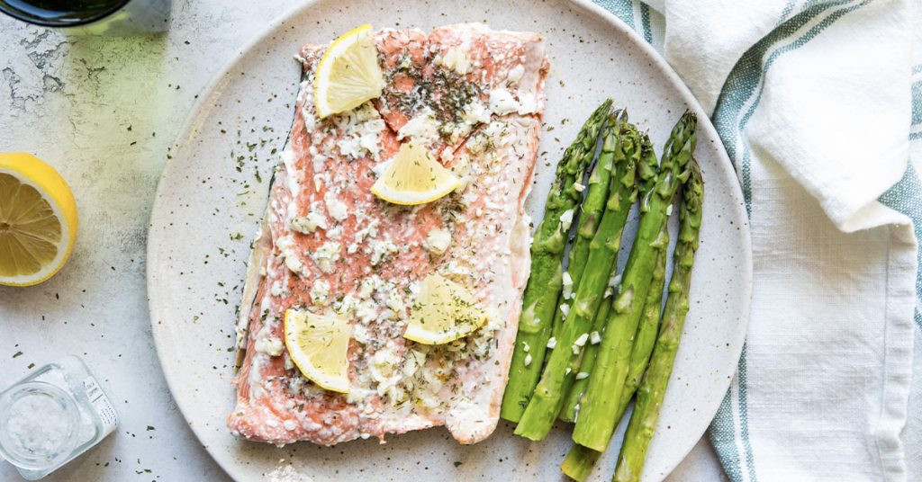 Salmon and asparagus is plated on a brown plate and topped with lemon.