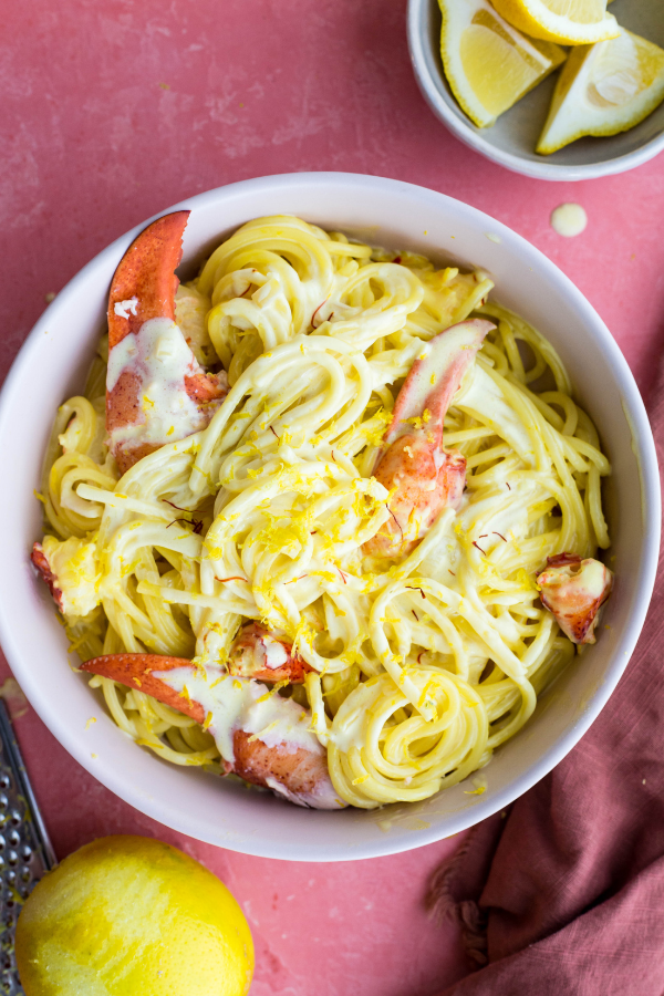 Lobster pasta is plated in a large white bowl next to sliced of lemon.