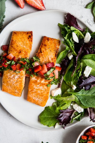 Salmon fillets are plated next to a salad and topped with fresh strawberry and basil relish.
