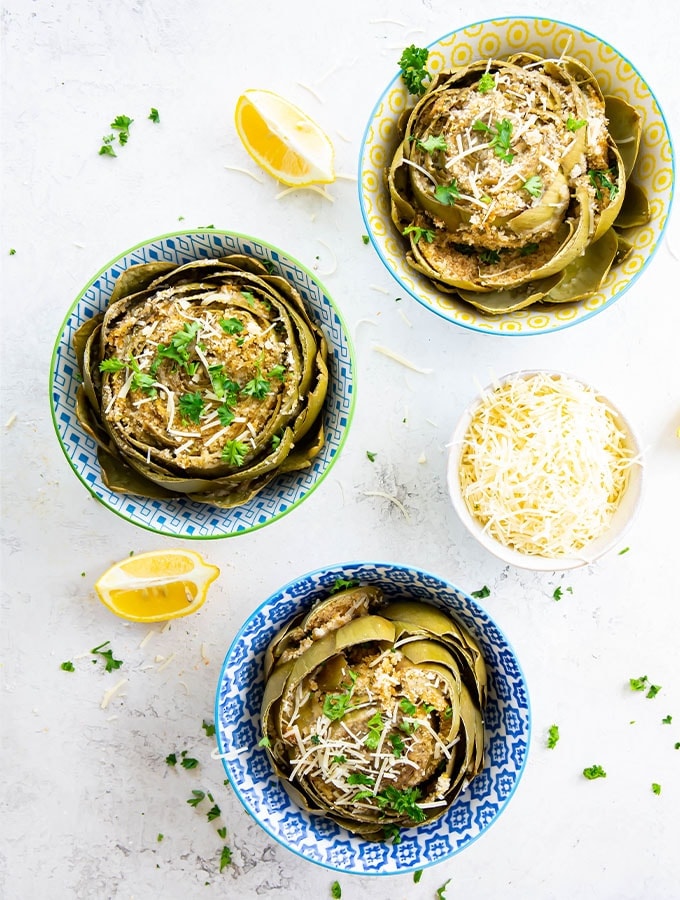 The artichokes are plated in separate bowls and plated next to sliced lemons and topped with cheese.