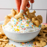 A hand is dunking an animal cracker into the dip that is plated in a white bowl.