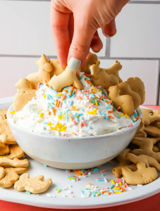 A hand is dunking an animal cracker into the dip that is plated in a white bowl.