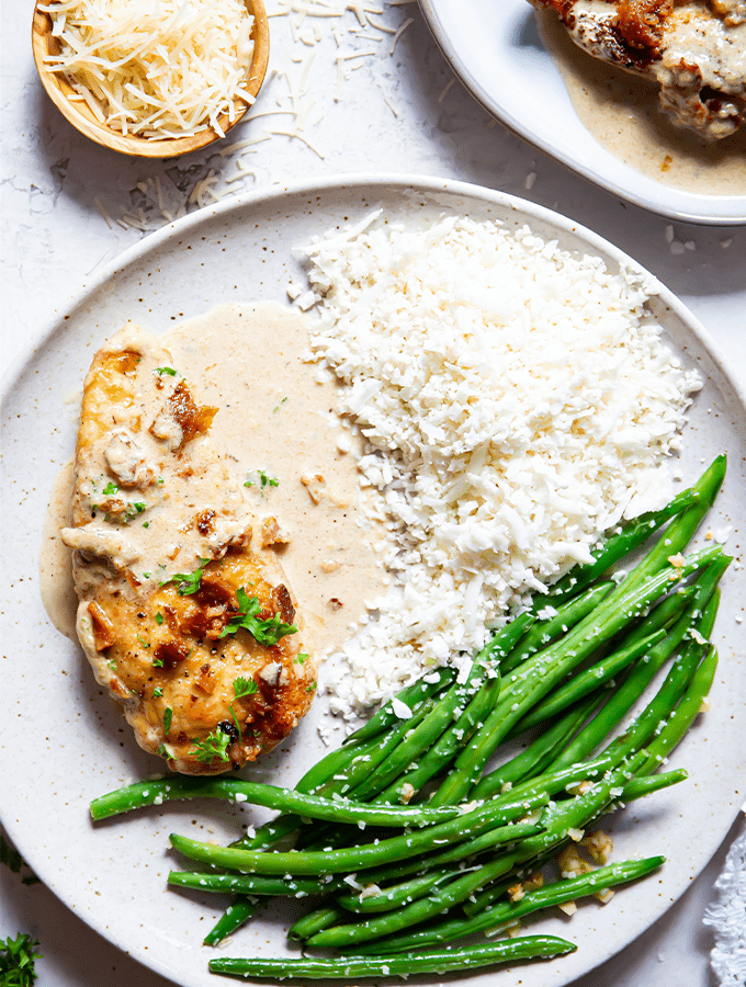 Chicken is plated with green beans and cauliflower rice, then topped with cream sauce.