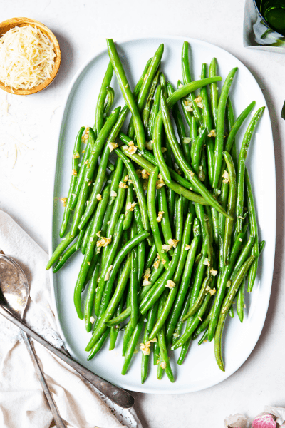 Green beans are plated on a white platter next to a small pinch bowl full of parmesan cheese.