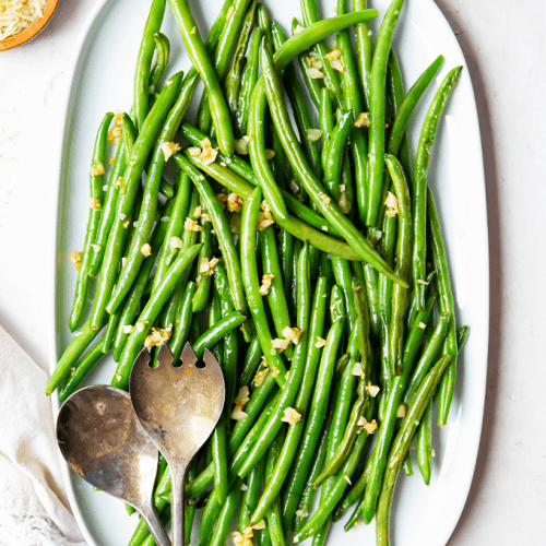 Green beans are plated on a white serving platter and topped with crispy garlic bits.