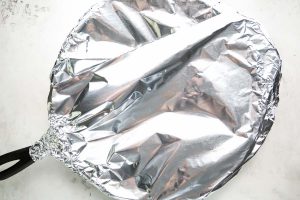 The chicken casserole in a cast iron pan is covered in tin foil before it is baked.