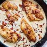 Crumbled bacon is sprinkled over the cream sauce and chicken filets in a cast iron pan.