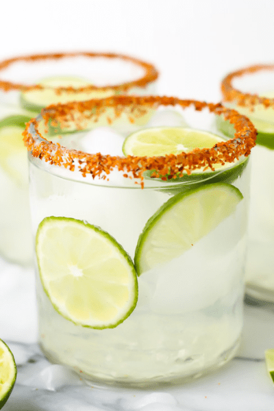A glass of margarita has Tajin around the tim and is topped with ice and wedge of lime.