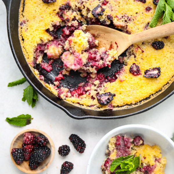 Blackberry cobbler is baked in a cast iron pan and served with a wooden spoon into a white bowl