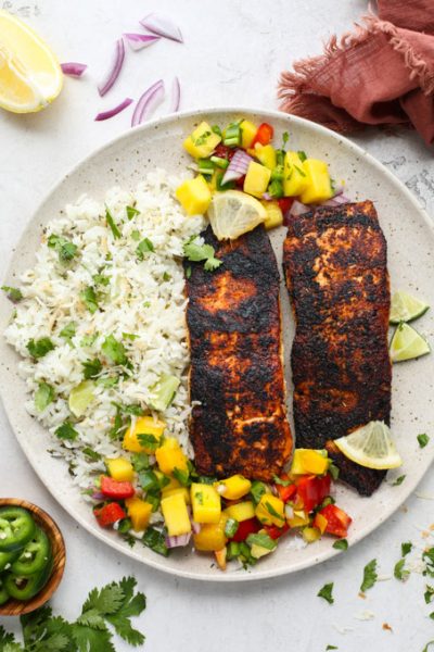 Blackened salmon is plated with coconut rice and mango salsa and topped with lemon wedges.