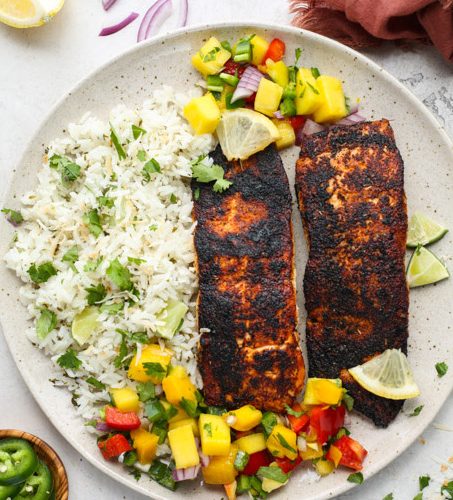 Blackened salmon is plated with coconut rice and mango salsa and topped with lemon wedges.