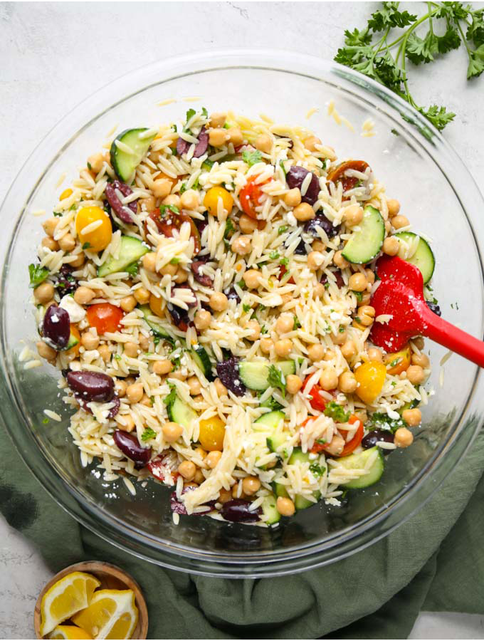 Orzo pasta salad is combined in a large bowl with a red spatula.