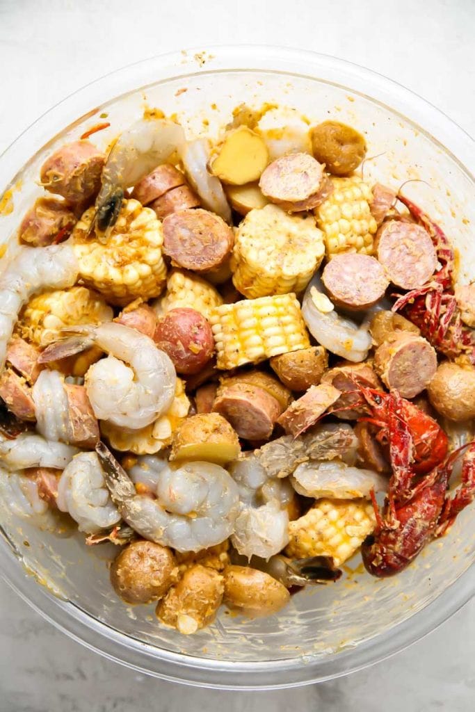 Shrimp, crawfish, sausage, potatoes, corn, butter, and Cajun seasoning is tossed together in a glass bowl.