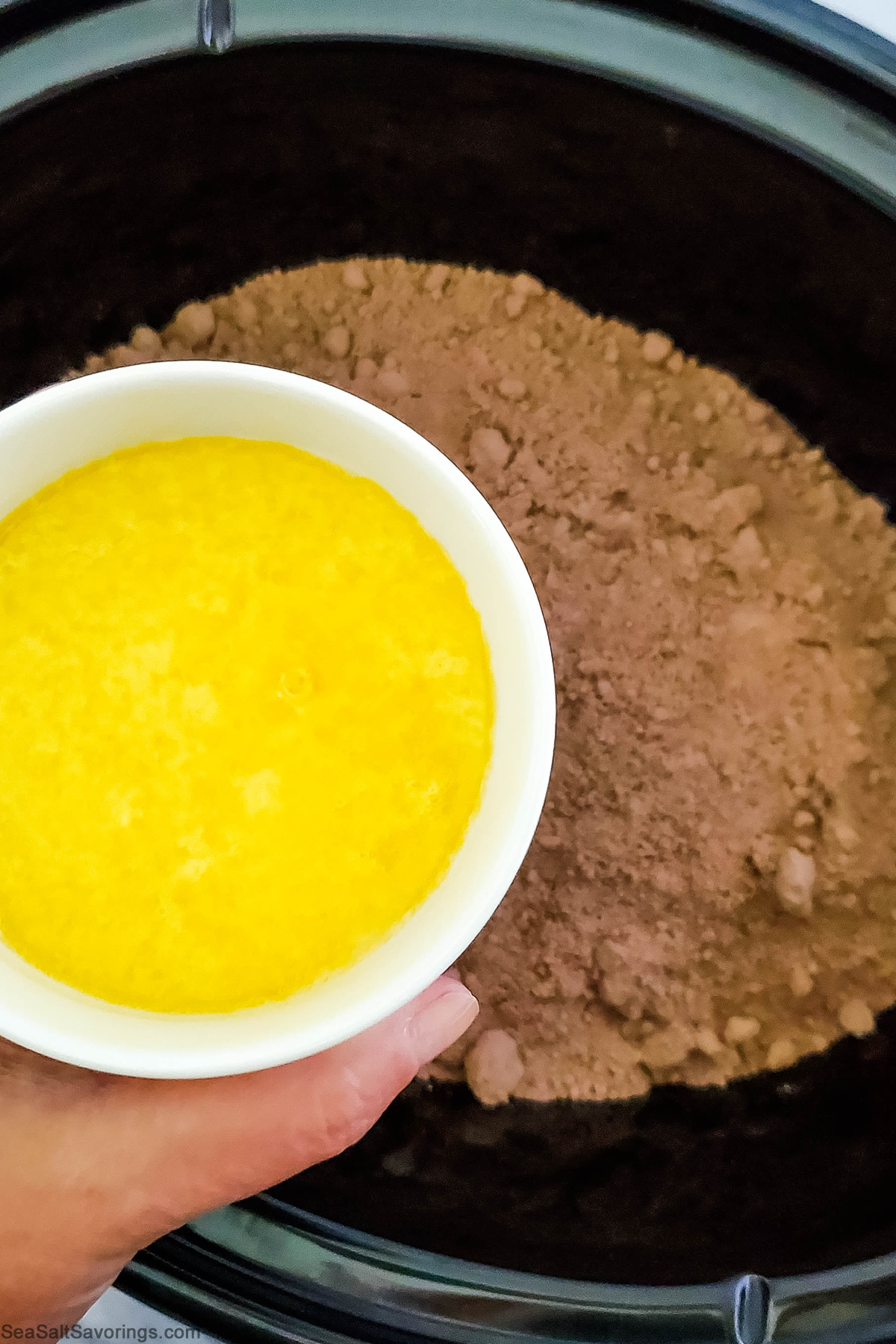 holding a cup of melted butter over the cake mix that has been spread on top of the cherry mixture