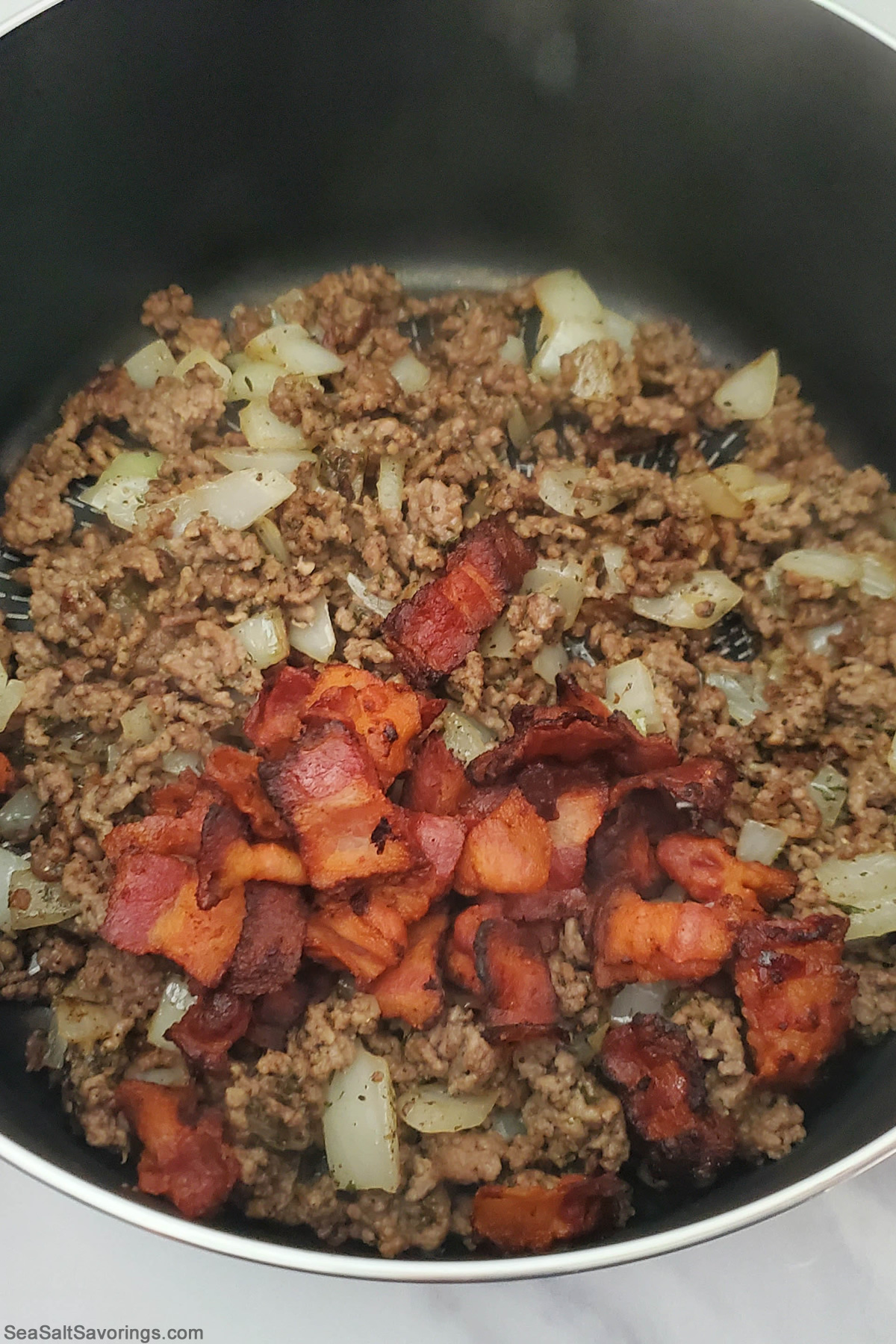 skillet with cooked beef and bacon and onions frying