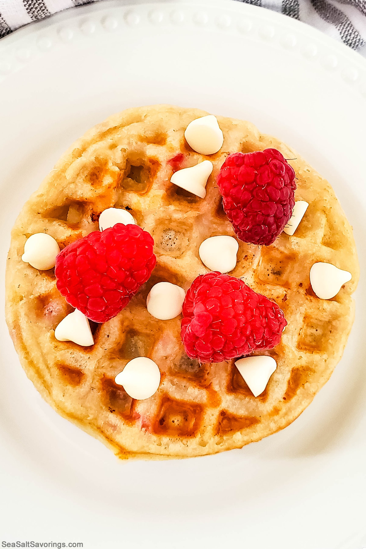 overhead view of stack of waffles on a plate with raspberries and white chocolate pieces sprinkled on top