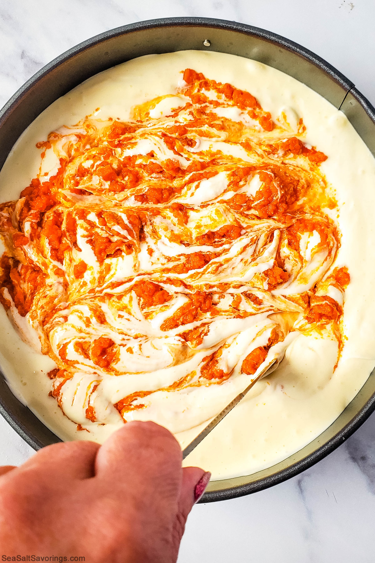 using a stick to swirl the pumpkin puree around the top of the cheesecake to create a swirl visual effect