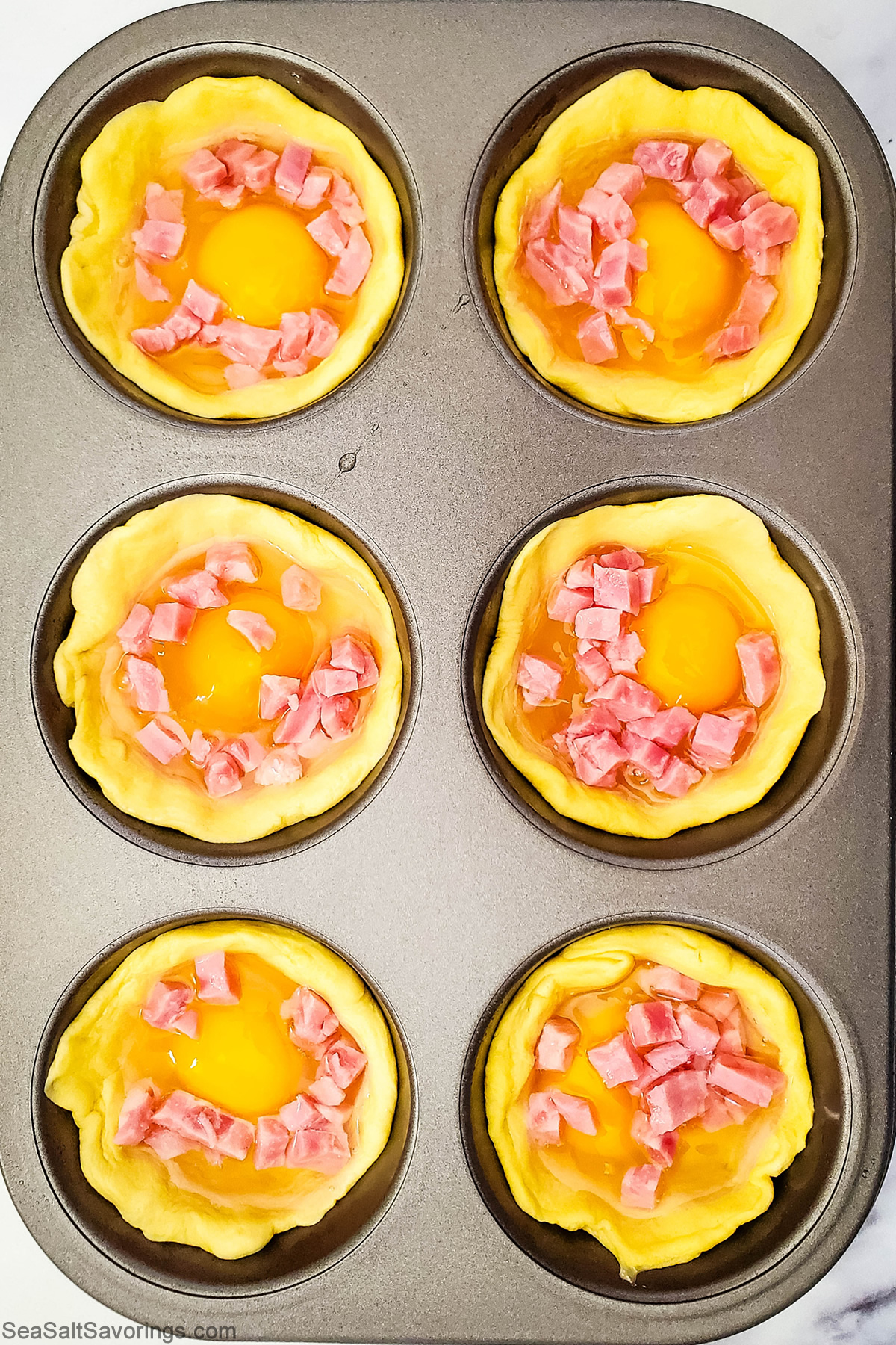 biscuits dough shaped into shells and filled with egg and ham in a muffin baking dish