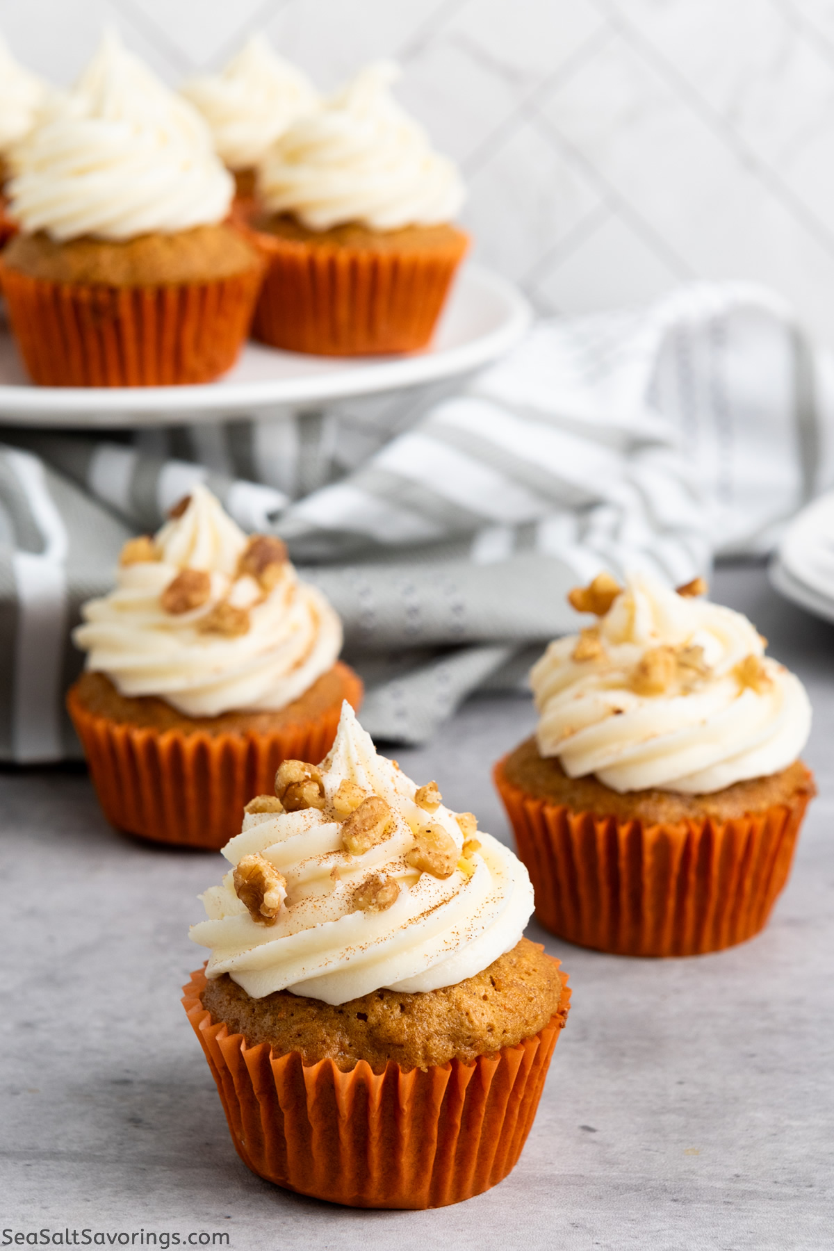 three carrot cupcakes with orange wrappers on a table with more cupcakes on a platter in the background
