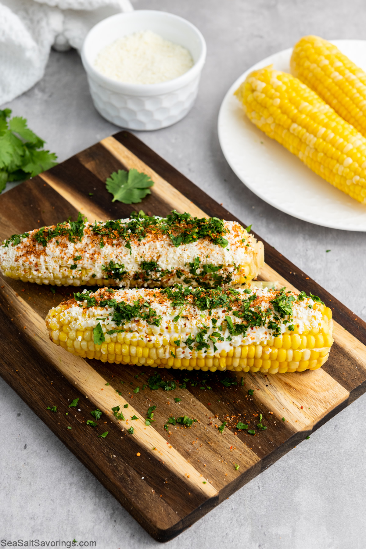 two corn on the cobs with seasons and toppings on a cutting board