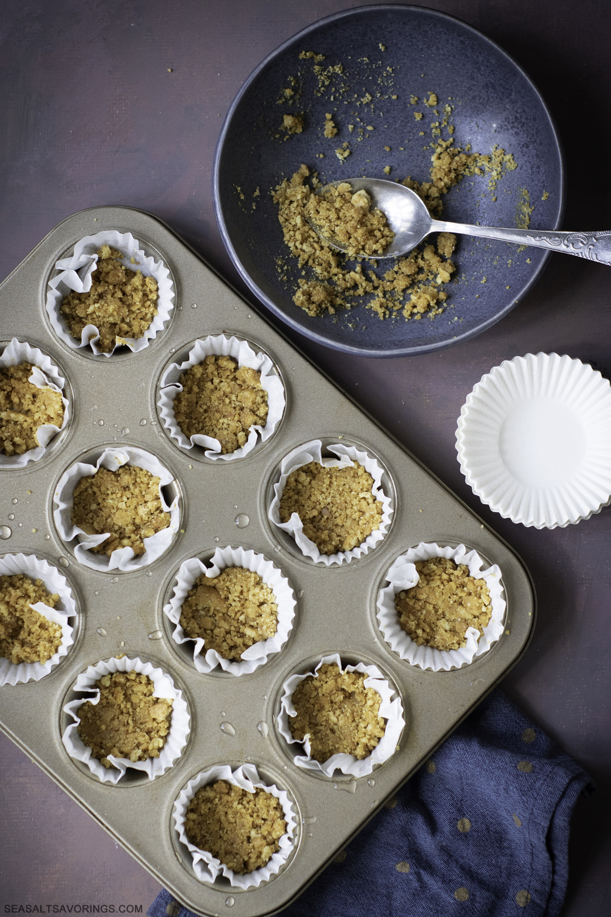 scooping mashed up graham cracker crumbs into cupcake liners in a cupcake baking pan