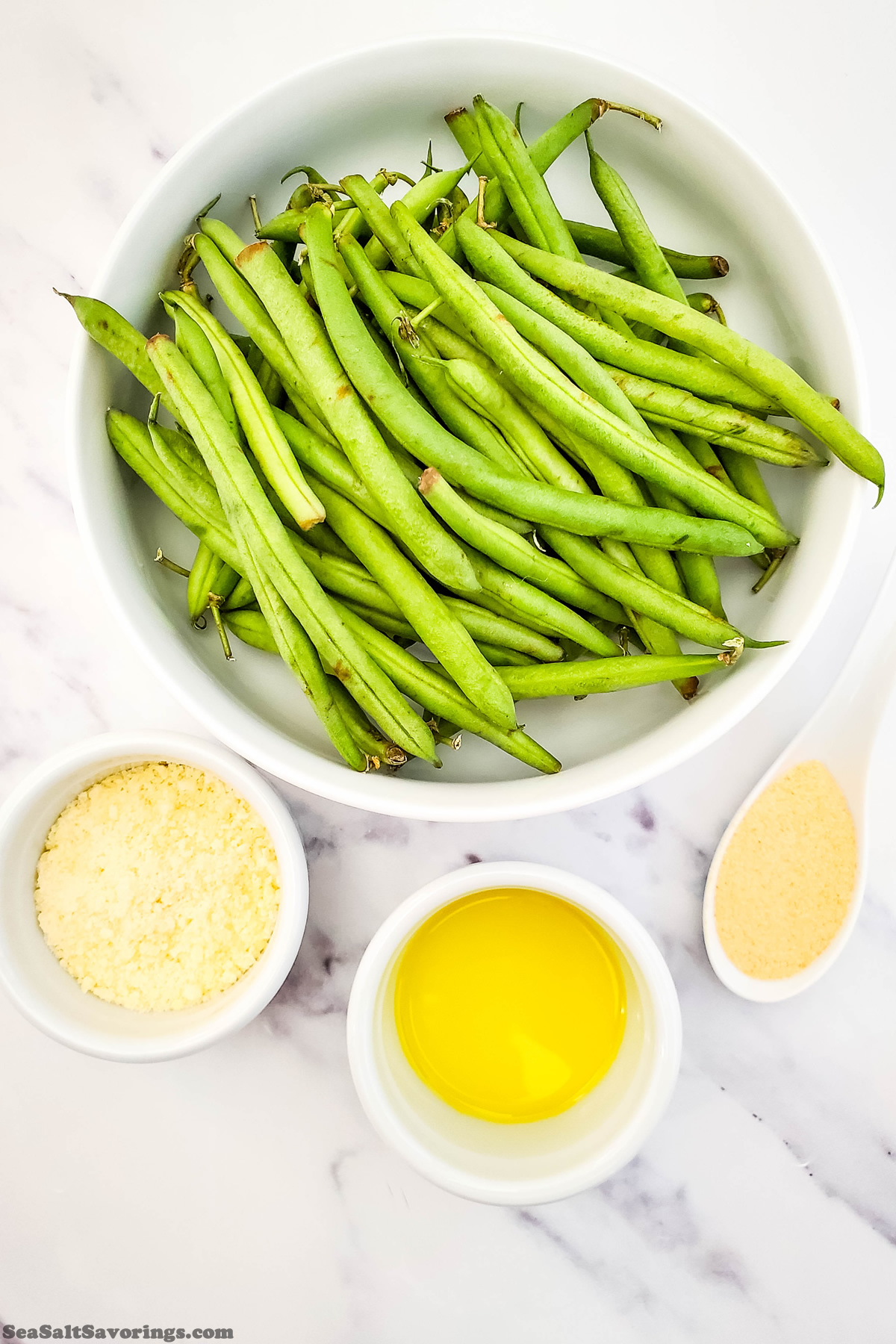 bowl of green beans next to smaller bowls of oil and parmesan cheese, the ingredients to make garlic parmesan green beans