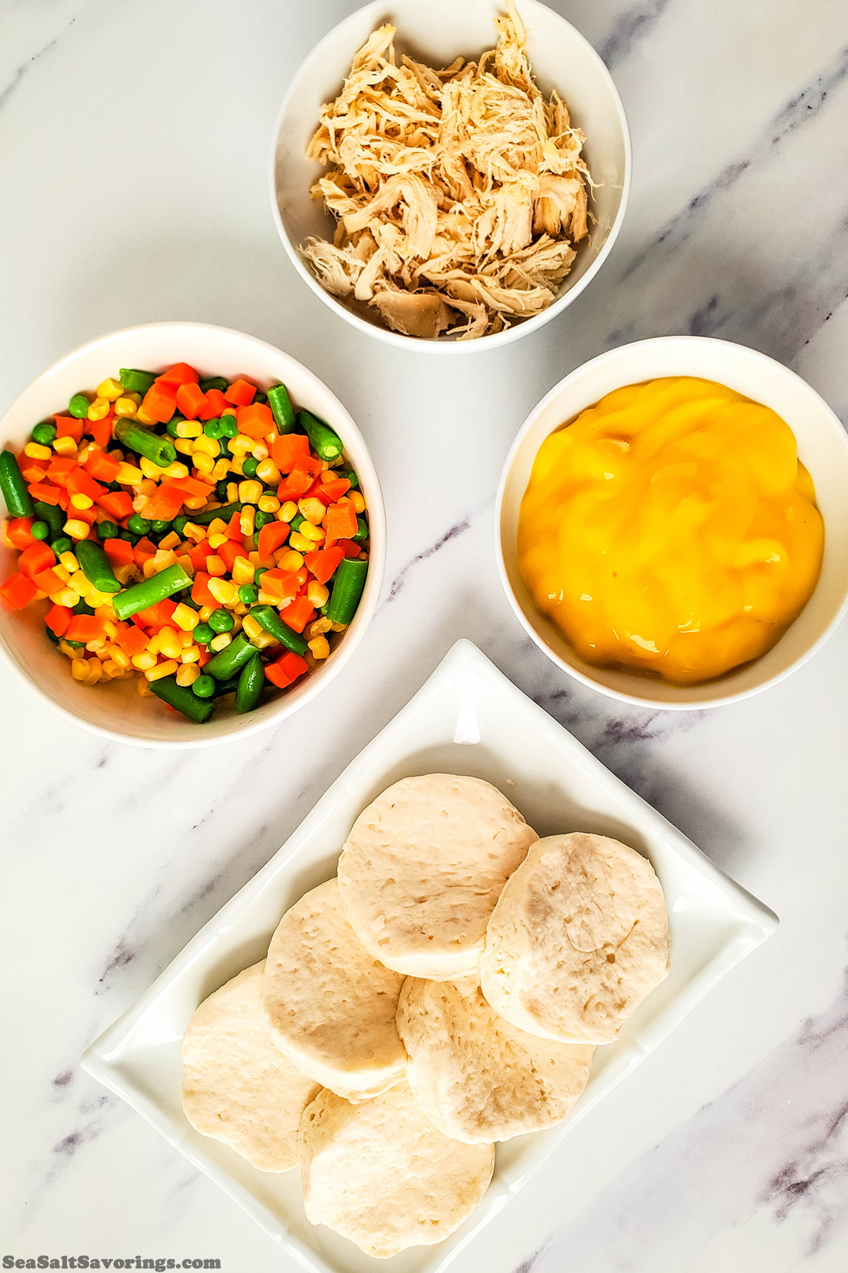 ingredients in bowls on a table including mixed vegetables, cheese, chicken and biscuits