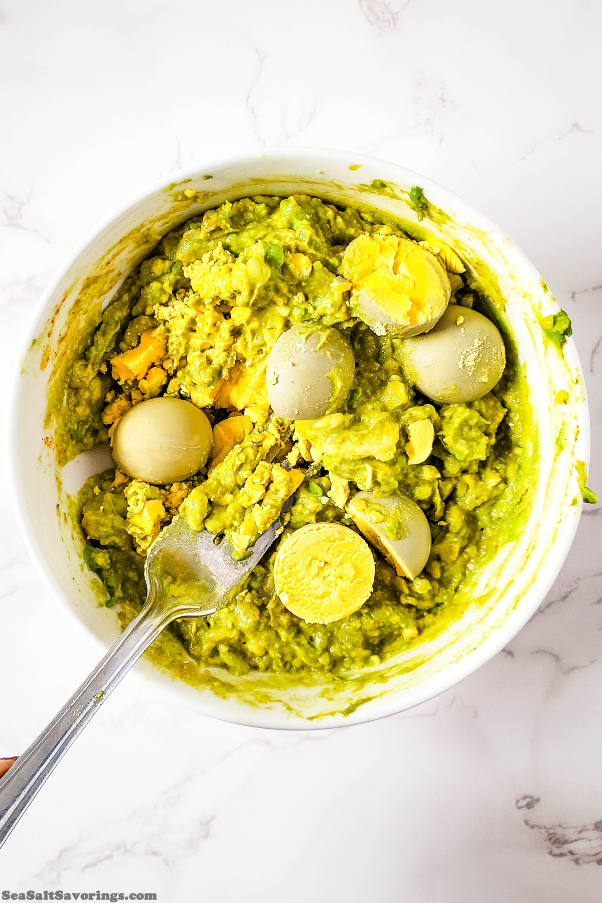 bowl of green mixture with egg yolks on top getting mashed with a fork